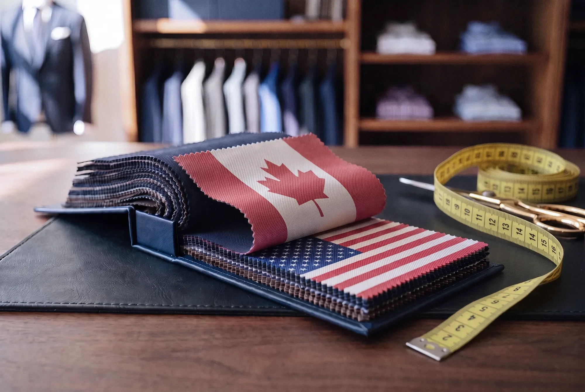 A tailor's fabric swatch book on a desk in a menswear store, with a Canadian flag swatch draped over an American flag swatch, a yellow tape measure and gold shears beside it